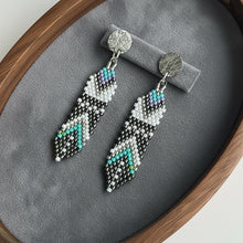 Load image into Gallery viewer, Pair of beaded earrings on a gray cushion inside a wooden frame.