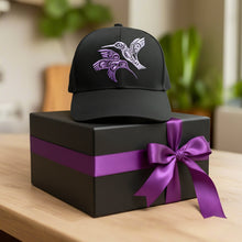 Load image into Gallery viewer, A black adjustable cap with a purple and white hummingbird design embroidered on the front, placed on a wooden surface.