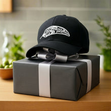 Load image into Gallery viewer, A black adjustable cap with a white embroidered design on the front, placed on a wooden surface with foliage in the background.