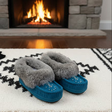 Load image into Gallery viewer, A pair of blue Laurentian Chief Moccasins with rabbit fur trim on a wooden surface with flowers in the background.