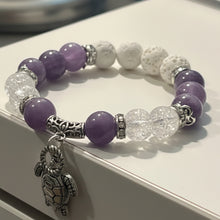 Load image into Gallery viewer, A beaded stretch bracelet with purple and white beads, clear faceted beads, and a silver turtle charm, resting on a white surface.