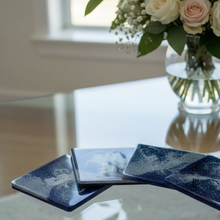 Load image into Gallery viewer, Set of blue ceramic coasters on a glass table with a vase of flowers in the background.