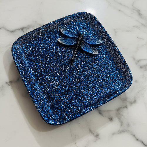 Blue glittery square object with a dragonfly on a beige surface