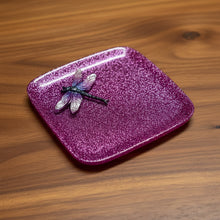 Load image into Gallery viewer, A square, glittery magenta-purple tray with rounded corners and a small dragonfly decoration on the top left, photographed on a light stone or tile surface.