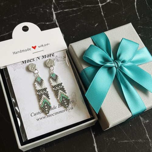 Mocs N More beaded earrings in packaging with a gift box featuring a turquoise bow on a dark surface.
