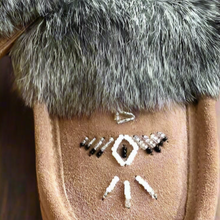 Load image into Gallery viewer, A pair of brown Laurentian Chief Moccasins with Thunderbird beading and rabbit fur lining displayed on a wooden surface with red roses around.