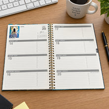 Load image into Gallery viewer, An open spiral-bound planner or calendar showing daily layout pages, placed on a desk in front of a black keyboard. The product pages are white with gray section headers and black text, and a small colorful photo insert is visible in the upper left.