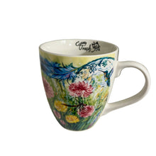 Load image into Gallery viewer, A white ceramic coffee mug with a colorful floral and bird illustration, featuring the brand name 'Touch Grass' printed inside the rim, displayed on a white shelf.