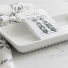 Load image into Gallery viewer, Earrings on a card with a white dish and patterned fabric on a marble surface