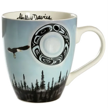 Load image into Gallery viewer, A ceramic coffee mug featuring a light blue sky design with a black and white totem circle, a flying eagle, and a black pine forest silhouette at the bottom. The brand name 'Anly Davis' is printed in black script on the inside of the mug.