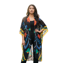 Load image into Gallery viewer, A woman modeling a black open-front kaftan-style cardigan with a vibrant multicolor floral and scroll pattern in red, yellow, blue, and green, set against a plain white background.